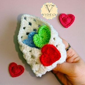 May include: A crocheted white pouch filled with blue, green, and red heart-shaped appliques. Two additional red heart appliques are placed near the pouch. The pouch has a square, open weave design. The image includes the text "VIDEO TUTORIAL".