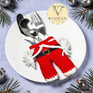 Könnte beinhalten: Ein Besteckhalter aus Häkelarbeit in Form einer Weihnachtsmannhose in Rot, Weiß und Schwarz mit einer goldfarbenen Gürtelschnalle. Der Halter enthält eine silberfarbene Gabel und ein Messer aus Metall auf einem weißen Teller.