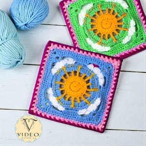 May include: Two crocheted squares with a sun and cloud design. The squares are blue and green with yellow suns and white clouds. The squares have a pink border. Blue yarn is in the background. The squares are handmade.