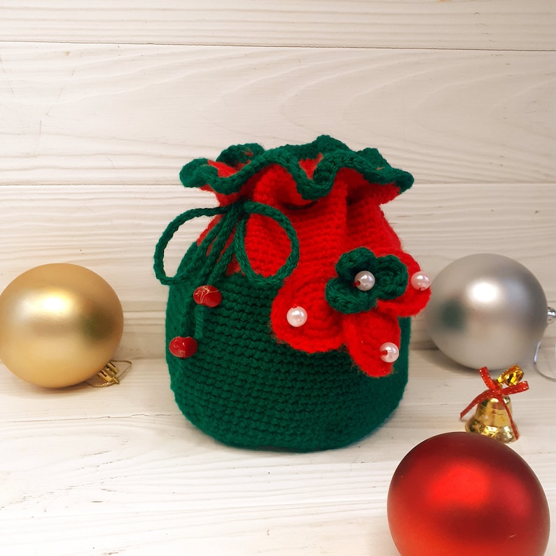 K&ouml;nnte beinhalten: Eine gr&uuml;ne und rote geh&auml;kelte Weihnachtsgeschenkt&uuml;te mit wei&szlig;en Perlen und roten Blumenakzenten.