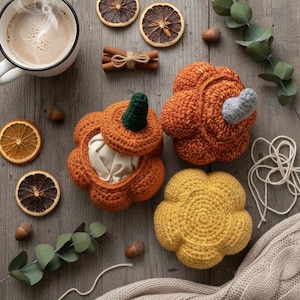 May include: An overhead shot features a cozy autumn scene. Crochet pumpkins in orange and yellow are arranged with dried orange slices, cinnamon sticks, acorns, and eucalyptus leaves. A mug of coffee and a cream-colored blanket complete the fall aesthetic.