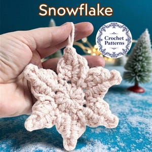 Könnte beinhalten: Hellrosa gehäkelter Schneeflocken-Ornament mit einer Schlaufe zum Aufhängen. Das Wort "Snowflake" steht oben im Bild. Das Ornament wird in einer Hand gehalten, mit einem kleinen Weihnachtsbaum im Hintergrund.