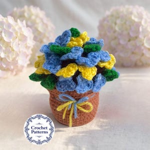 Puede incluir: Un arreglo floral de ganchillo en una maceta marrón, con flores azules, amarillas y verdes. La maceta está atada con un lazo azul y amarillo. La imagen incluye un logotipo que dice "Crochet Patterns".