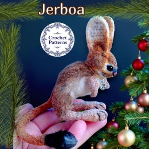 Puede incluir: Una figura de jerbo hecha a mano, con pelaje marrón y crema, orejas grandes y una cola larga. El jerbo tiene un pequeño colgante y se sostiene en una mano. La imagen incluye el texto "Jerboa" y "Crochet Patterns".