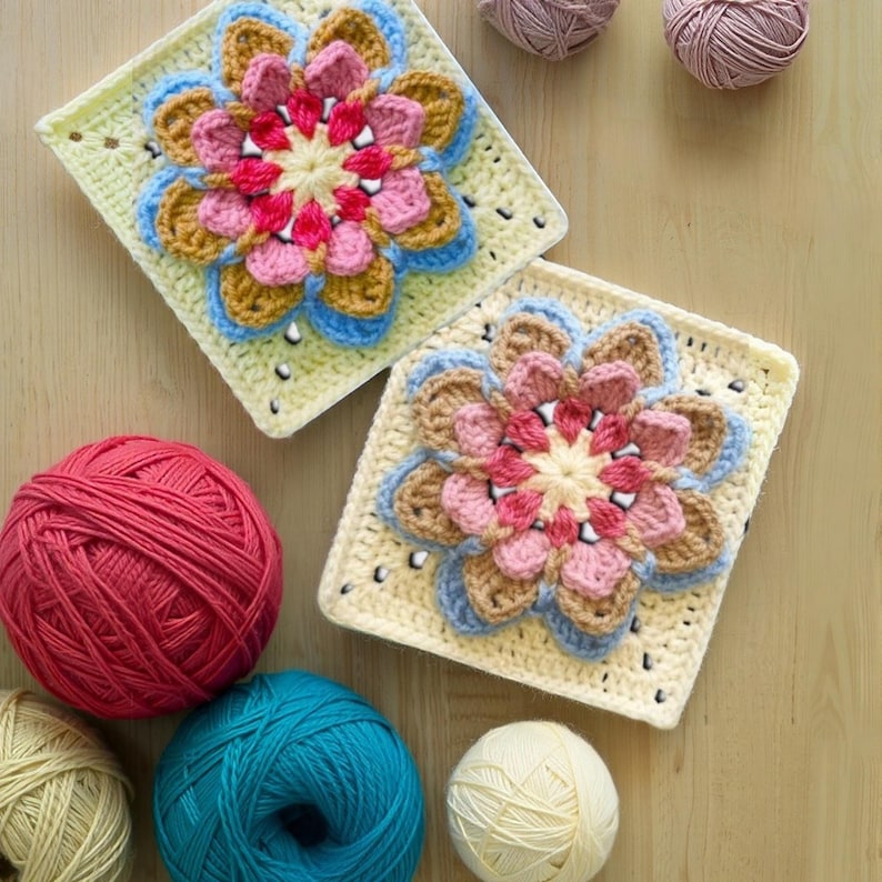 May include: Two crocheted flower squares with a cream background, featuring pink, blue, and brown petals. Several balls of yarn in pink, blue, and cream are scattered around the squares, suggesting a crafting project.