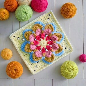May include: A handmade crochet square with a floral design in shades of pink, blue, yellow, and brown. Several balls of yarn in various colors, including orange, green, and pink, are scattered around the square on a white wooden surface. The square is approximately 8 inches by 8 inches.