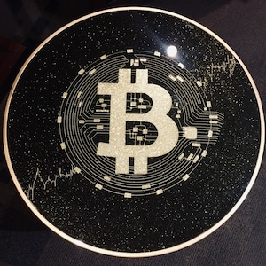 Op de afbeelding: Een zwarte ronde tafelblad met een gouden glitterafwerking. Het tafelblad heeft een groot, wit, gestileerd Bitcoin-symbool met circuitboard-achtige patronen eromheen.
