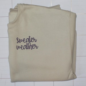 Pu&ograve; includere: Una felpa beige chiaro con il testo "sweater weather" ricamato in viola sul davanti.