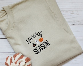 May include: A beige sweatshirt with the text "spooky season" embroidered in black. The text is accompanied by a small embroidered ghost, witch's hat, and pumpkin.