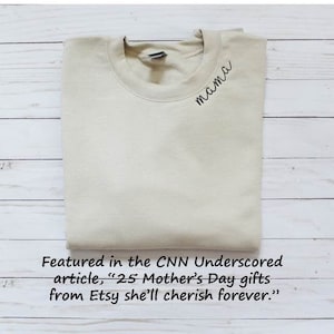 May include: A light beige sweatshirt with the word "mama" embroidered on the front. The sweatshirt is folded and photographed on a white wooden surface. The text "Featured in the CNN Underscored article, "25 Mother's Day gifts from Etsy she'll cherish forever."