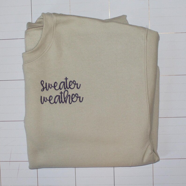 Pu&ograve; includere: Una felpa beige chiaro con il testo "sweater weather" ricamato in viola sul davanti.