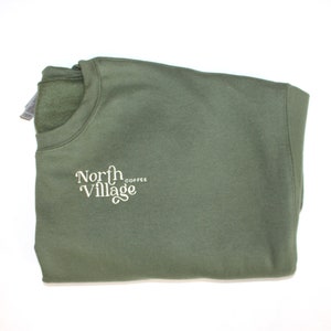 May include: An olive green sweatshirt with the words "North Village Coffee" in a white, stylized font. The sweatshirt is folded neatly, showcasing the crew neck and the embroidered logo on the front.