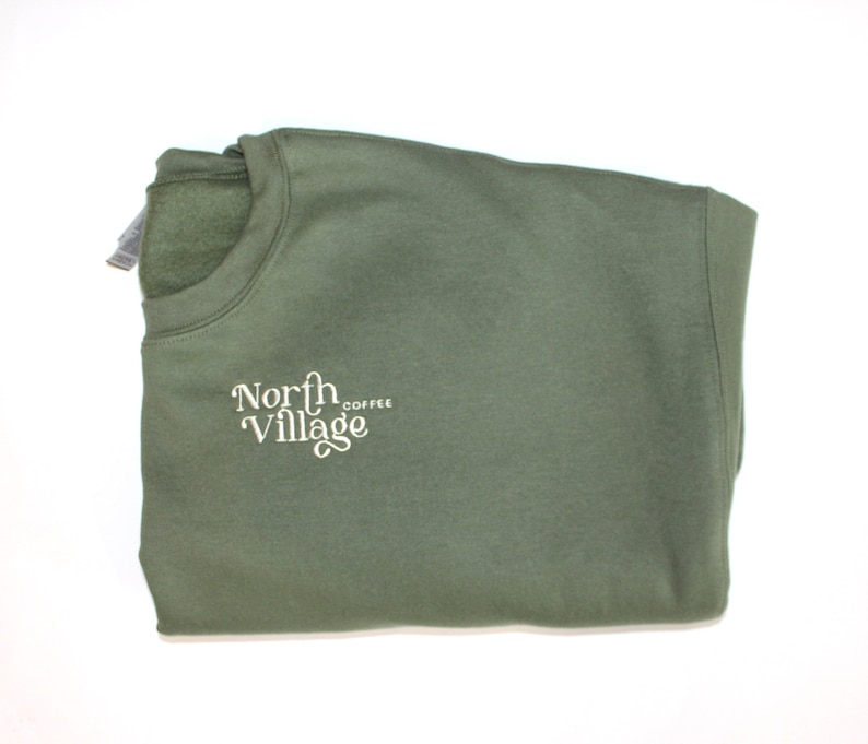 May include: An olive green sweatshirt with the words "North Village Coffee" in a white, stylized font. The sweatshirt is folded neatly, showcasing the crew neck and the embroidered logo on the front.