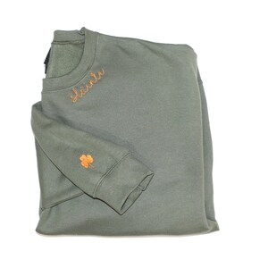 May include: A green sweatshirt with the word "Sl&aacute;inte" embroidered on the front in orange thread. A small orange shamrock is embroidered on the left sleeve.