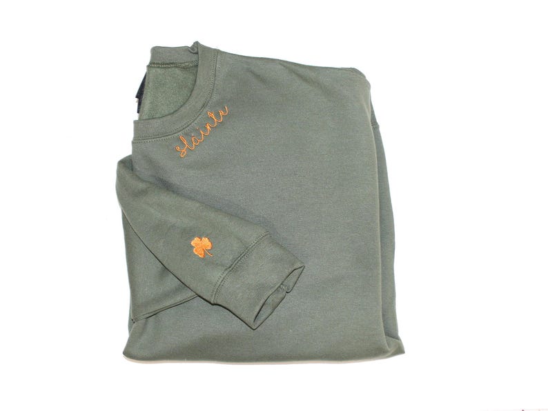 May include: A green sweatshirt with the word "Sl&aacute;inte" embroidered on the front in orange thread. A small orange shamrock is embroidered on the left sleeve.