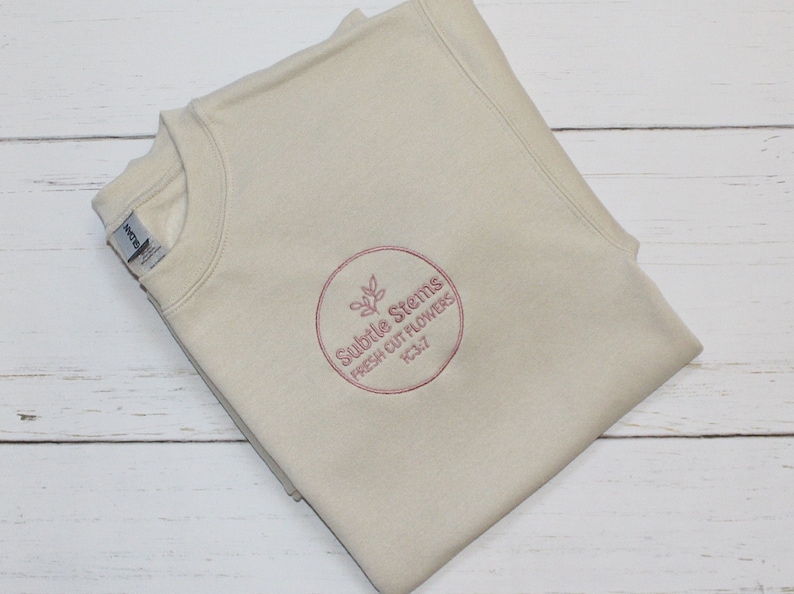 May include: A beige crewneck sweatshirt with a circular embroidered logo. The logo features the text "Subtle Stems Fresh Cut Flowers" in a burgundy color, along with a floral design. The sweatshirt is folded and appears to be made of a soft material.