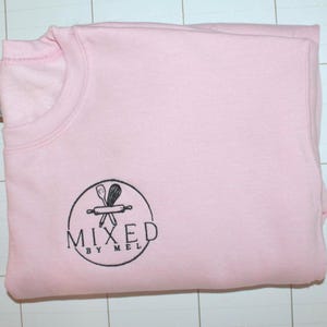 Pu&ograve; includere: Una felpa rosa chiaro piegata con scollo tondo. La felpa ha un logo circolare nero con la scritta "MIXED BY MEL" e una grafica di un mattarello e utensili.