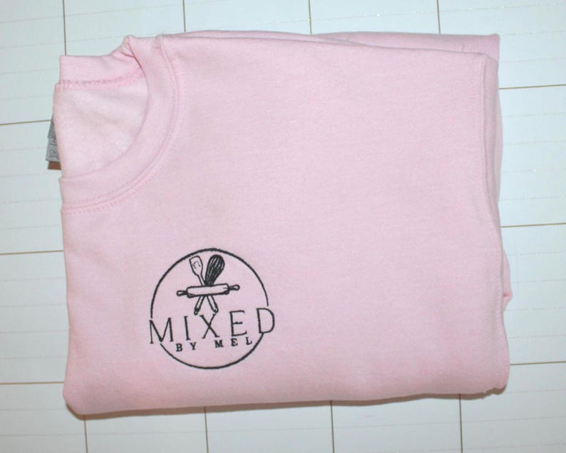 Pu&ograve; includere: Una felpa rosa chiaro piegata con scollo tondo. La felpa ha un logo circolare nero con la scritta "MIXED BY MEL" e una grafica di un mattarello e utensili.