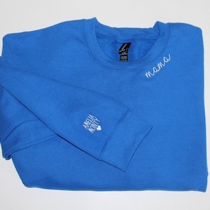 Puede incluir: Una sudadera azul real con la palabra "mama" bordada en blanco en el pecho. La manga izquierda tiene las palabras "Amelia Monty" bordadas en blanco. La sudadera est&aacute; doblada sobre una superficie blanca.