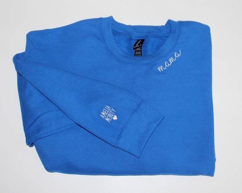 Puede incluir: Una sudadera azul real con la palabra "mama" bordada en blanco en el pecho. La manga izquierda tiene las palabras "Amelia Monty" bordadas en blanco. La sudadera est&aacute; doblada sobre una superficie blanca.