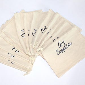 May include: A stack of 12 natural canvas zippered pouches with black embroidery. The pouches are labeled "Art" and "Art Supplies".