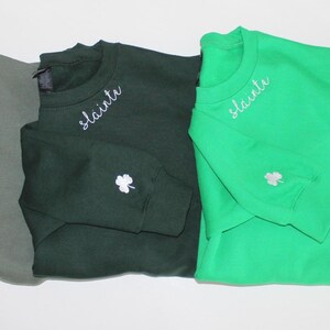 May include: Three sweatshirts in different shades of green with the word "slainte" embroidered on the necklines. Each sweatshirt has a white shamrock embroidered on the sleeve.