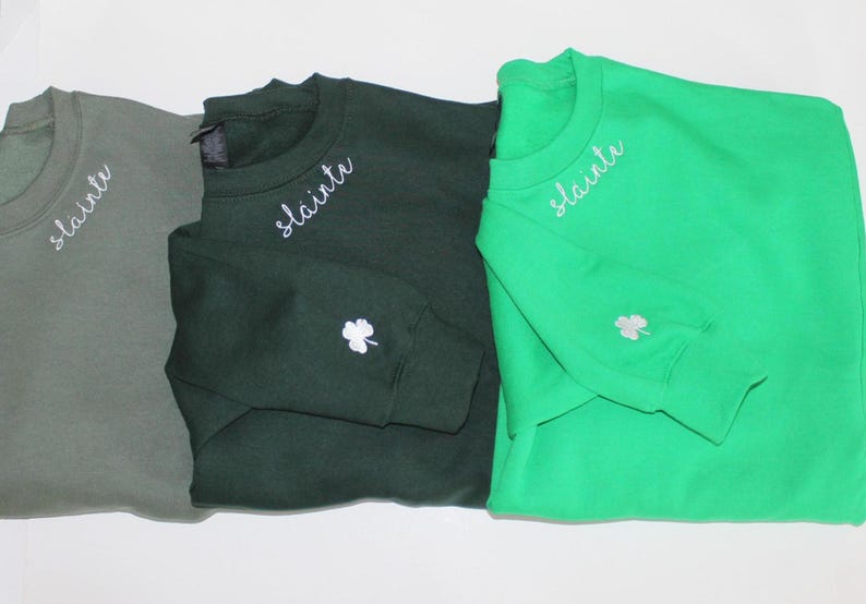 May include: Three sweatshirts in different shades of green with the word "slainte" embroidered on the necklines. Each sweatshirt has a white shamrock embroidered on the sleeve.