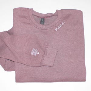 Puede incluir: Una sudadera de color malva con cuello redondo y la palabra "mama" bordada en blanco en el pecho. El pu&ntilde;o de la manga izquierda lleva el texto "Graham Audrey" y un peque&ntilde;o coraz&oacute;n. La sudadera est&aacute; doblada cuidadosamente.