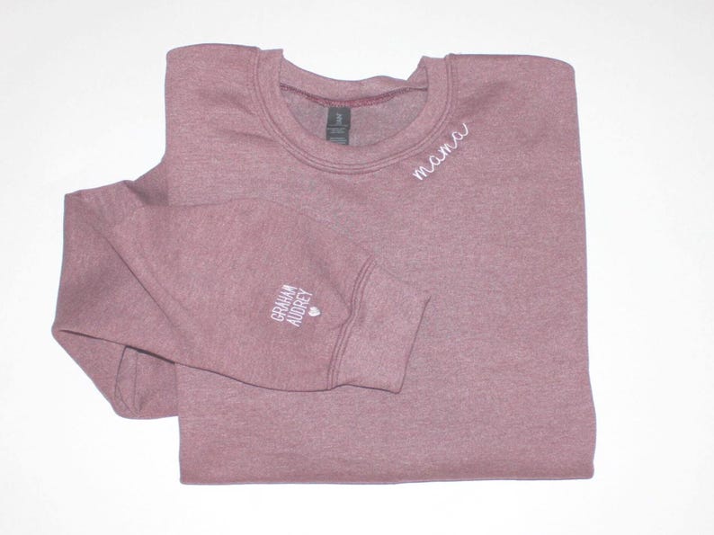 Puede incluir: Una sudadera de color malva con cuello redondo y la palabra "mama" bordada en blanco en el pecho. El pu&ntilde;o de la manga izquierda lleva el texto "Graham Audrey" y un peque&ntilde;o coraz&oacute;n. La sudadera est&aacute; doblada cuidadosamente.