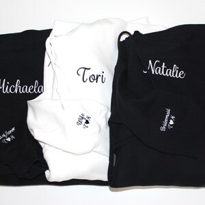 May include: Black and white hoodies with custom embroidered names and titles. The black hoodies have the names Michaela and Natalie, with "Maid of Honor" and "Bridesmaid" on the sleeves. The white hoodie has the name "Tori" and "Wife" on the sleeve.