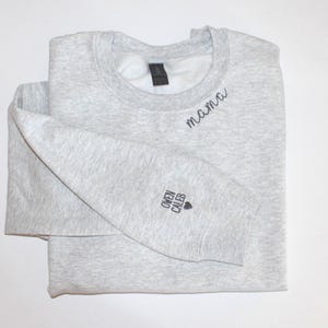 Puede incluir: Una sudadera gris claro con la palabra "mama" bordada en escritura negra en el pecho. La manga izquierda presenta los nombres "Owen Caleb" y un s&iacute;mbolo de coraz&oacute;n. La sudadera est&aacute; doblada.