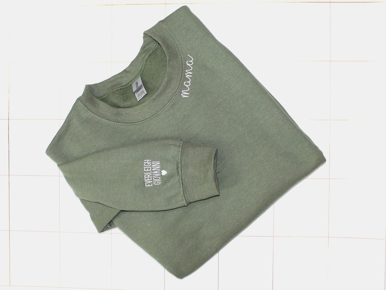 Puede incluir: Sudadera verde oliva con la palabra "mama" en escritura blanca en la parte superior del pecho. La manga izquierda presenta los nombres "EVERLEIGH GIOVANNI" en letras blancas, con un peque&ntilde;o s&iacute;mbolo de coraz&oacute;n. La sudadera es de material suave.