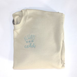 Pode incluir: Uma camisola bege com o texto "ALWAYS cold" bordado a azul na frente. O texto &eacute; rodeado por tr&ecirc;s pequenos desenhos de flocos de neve.