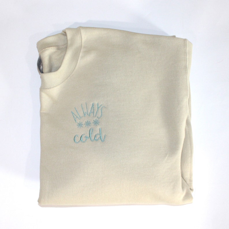 Pode incluir: Uma camisola bege com o texto "ALWAYS cold" bordado a azul na frente. O texto &eacute; rodeado por tr&ecirc;s pequenos desenhos de flocos de neve.