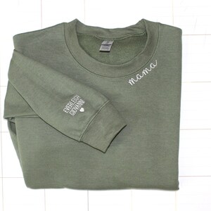 Puede incluir: Sudadera verde oliva con la palabra "mama" bordada en blanco en el pecho. La manga izquierda presenta los nombres bordados "Everleigh Giovanni" y un peque&ntilde;o coraz&oacute;n blanco. Una prenda superior informal y c&oacute;moda.