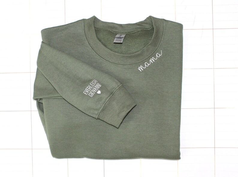 Puede incluir: Sudadera verde oliva con la palabra "mama" bordada en blanco en el pecho. La manga izquierda presenta los nombres bordados "Everleigh Giovanni" y un peque&ntilde;o coraz&oacute;n blanco. Una prenda superior informal y c&oacute;moda.