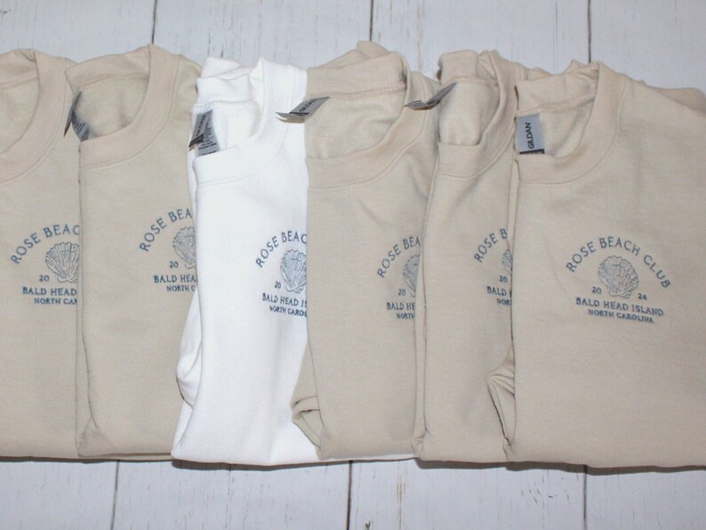 Pu&ograve; includere: Una collezione di felpe girocollo piegate in vari colori neutri, tra cui beige e bianco. Ogni felpa presenta una grafica blu con il testo "ROSE BEACH CLUB 24 BALD HEAD ISLAND NORTH CAROLINA".