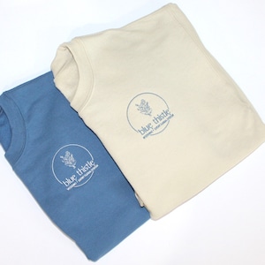 May include: Two folded sweatshirts, one in a muted blue and the other in a cream color. Each has a circular logo with a floral design and the text "blue thistle" and "wedding + event floral design" in a matching blue.