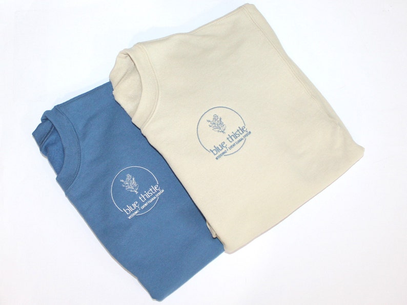 May include: Two folded sweatshirts, one in a muted blue and the other in a cream color. Each has a circular logo with a floral design and the text "blue thistle" and "wedding + event floral design" in a matching blue.