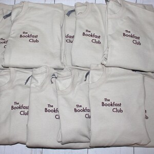 May include: A collection of beige crew neck t-shirts, each folded and displaying the embroidered text "The Bookfast Club" in a dark brown color. The shirts are neatly stacked on a light-colored surface.