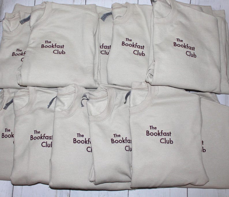 May include: A collection of beige crew neck t-shirts, each folded and displaying the embroidered text "The Bookfast Club" in a dark brown color. The shirts are neatly stacked on a light-colored surface.