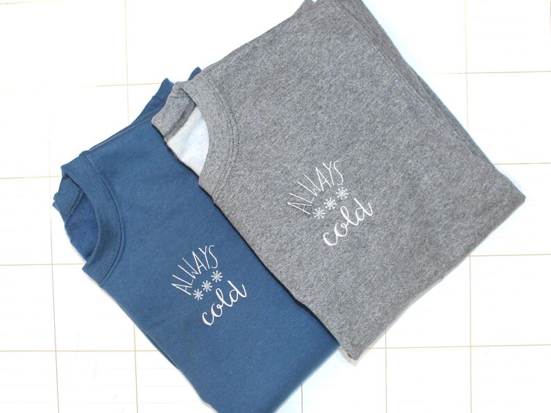Pode incluir: Duas sweatshirts com o texto "Always cold" bordado. Uma sweatshirt &eacute; azul e a outra &eacute; cinza.