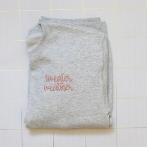 Pu&ograve; includere: Una felpa grigia con il testo "sweater weather" ricamato in rosa sul davanti.