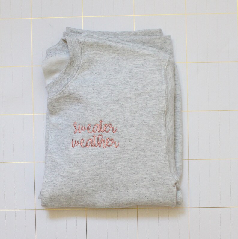 Pu&ograve; includere: Una felpa grigia con il testo "sweater weather" ricamato in rosa sul davanti.