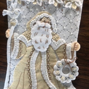 Puede incluir: Una media de Navidad de fieltro blanco con una figura de Pap&aacute; Noel beige que sostiene un bast&oacute;n y una corona. La media est&aacute; decorada con cuentas blancas y plateadas y flores bordadas.