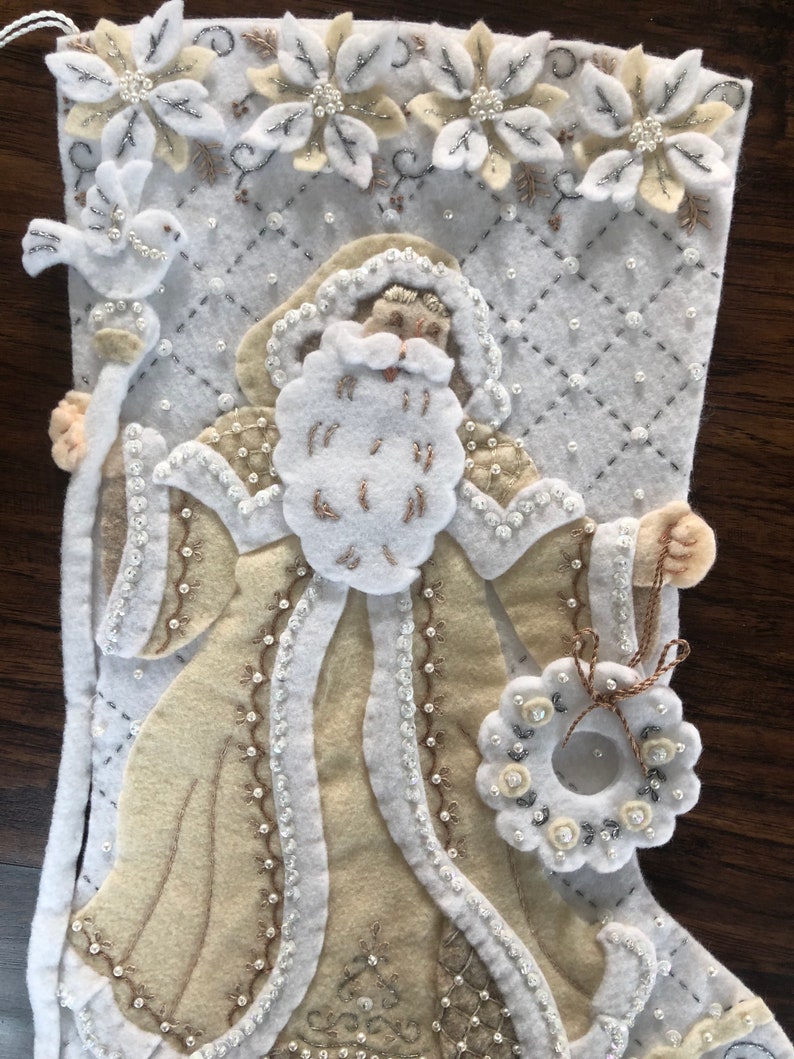 Puede incluir: Una media de Navidad de fieltro blanco con una figura de Pap&aacute; Noel beige que sostiene un bast&oacute;n y una corona. La media est&aacute; decorada con cuentas blancas y plateadas y flores bordadas.