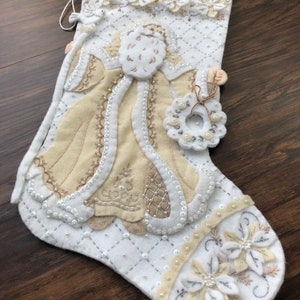 Puede incluir: Calcet&iacute;n de Navidad de fieltro blanco con un Pap&aacute; Noel bordado en beige que sostiene una corona. El calcet&iacute;n est&aacute; decorado con cuentas y flores blancas y beige.