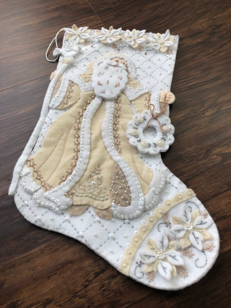 Puede incluir: Calcet&iacute;n de Navidad de fieltro blanco con un Pap&aacute; Noel bordado en beige que sostiene una corona. El calcet&iacute;n est&aacute; decorado con cuentas y flores blancas y beige.