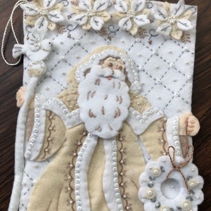 Puede incluir: Una media de Navidad de fieltro blanco con una figura de Pap&aacute; Noel beige que sostiene una corona. La media est&aacute; decorada con cuentas blancas y plateadas, lentejuelas y flores bordadas.