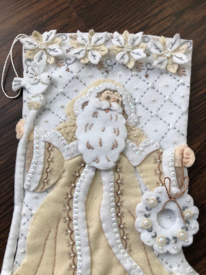 Puede incluir: Una media de Navidad de fieltro blanco con una figura de Pap&aacute; Noel beige que sostiene una corona. La media est&aacute; decorada con cuentas blancas y plateadas, lentejuelas y flores bordadas.